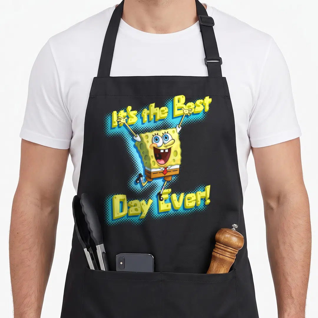 Squarepant general apron - SpongeBob jumping between arched yellow text It's the Best Day Ever on blue halftone — shown on a black cotton canvas apron against a white background