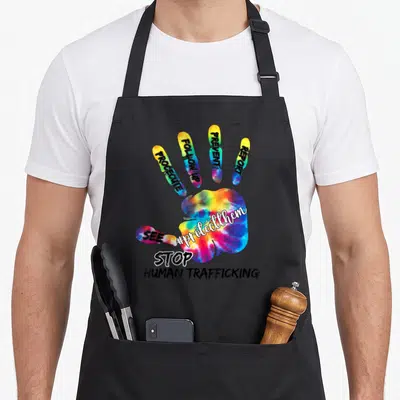 Raise human trafficking awareness apron - Tie-dye hand with PROSECUTE, REPORT, STOP HUMAN TRAFFICKING text on fingers — shown on a black cotton canvas apron against a white background