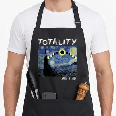 Solar april apron - Totality April 8 2024 text above blue yellow watercolor eclipse arc over cypress tree village — shown on a black cotton canvas apron against a white background