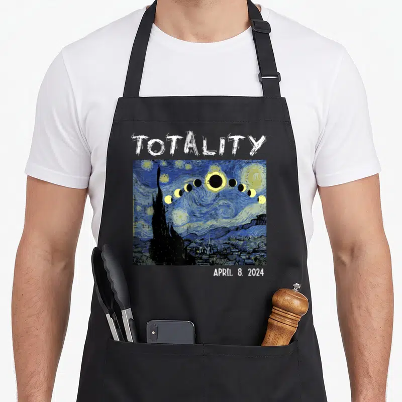 Solar april apron - Totality April 8 2024 text above blue yellow watercolor eclipse arc over cypress tree village — shown on a black cotton canvas apron against a white background