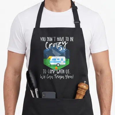 Camp us aprons - Camper and tent in hexagon with text You Don't Have To Be Crazy To Camp With Us — shown on a black cotton canvas apron against a white background