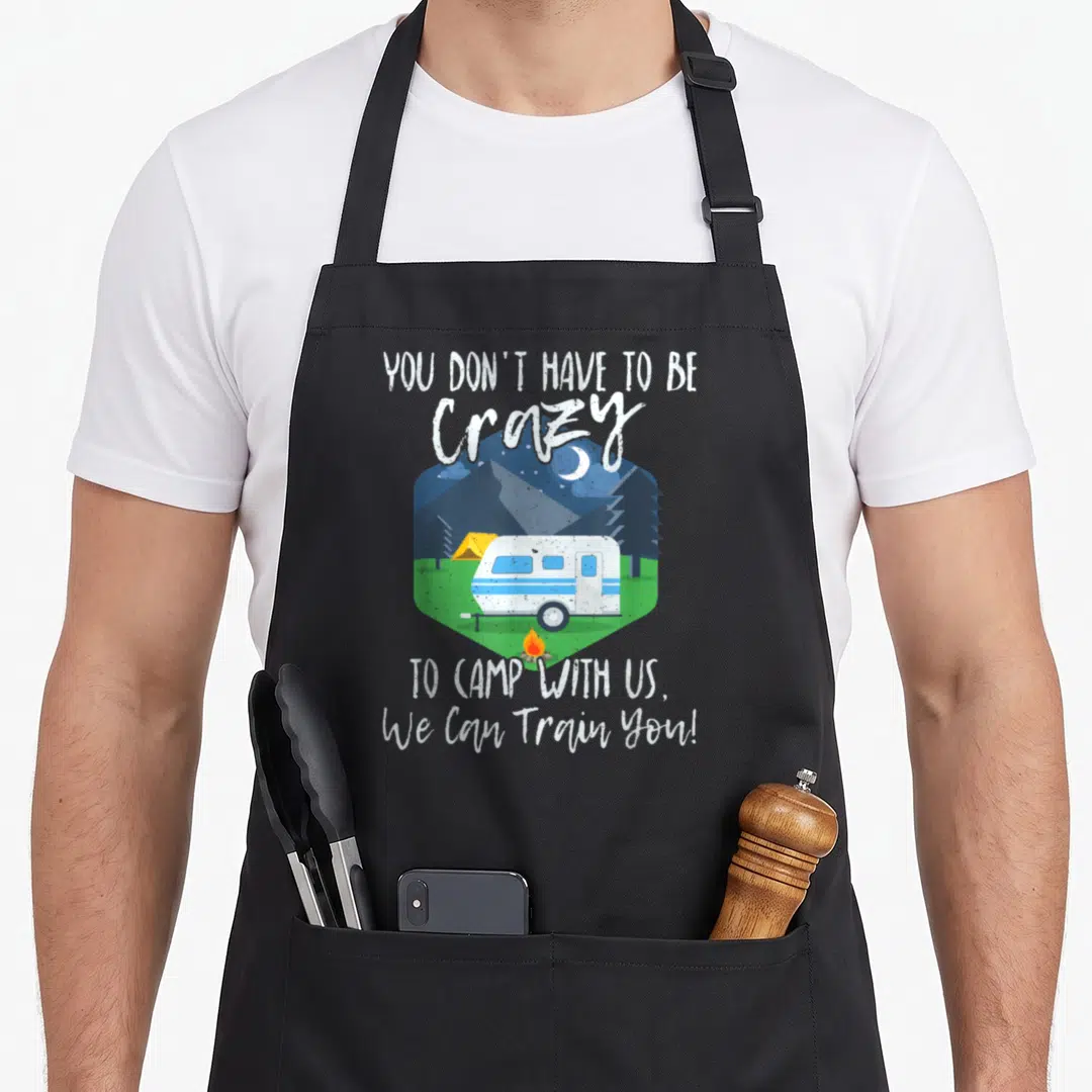 Camp us aprons - Camper and tent in hexagon with text You Don't Have To Be Crazy To Camp With Us — shown on a black cotton canvas apron against a white background