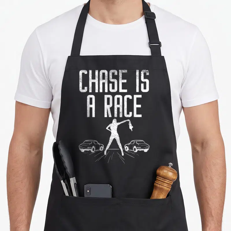 Chase car apron - White text chase is a race above woman holding checkered flag between two cars — shown on a black cotton canvas apron against a white background