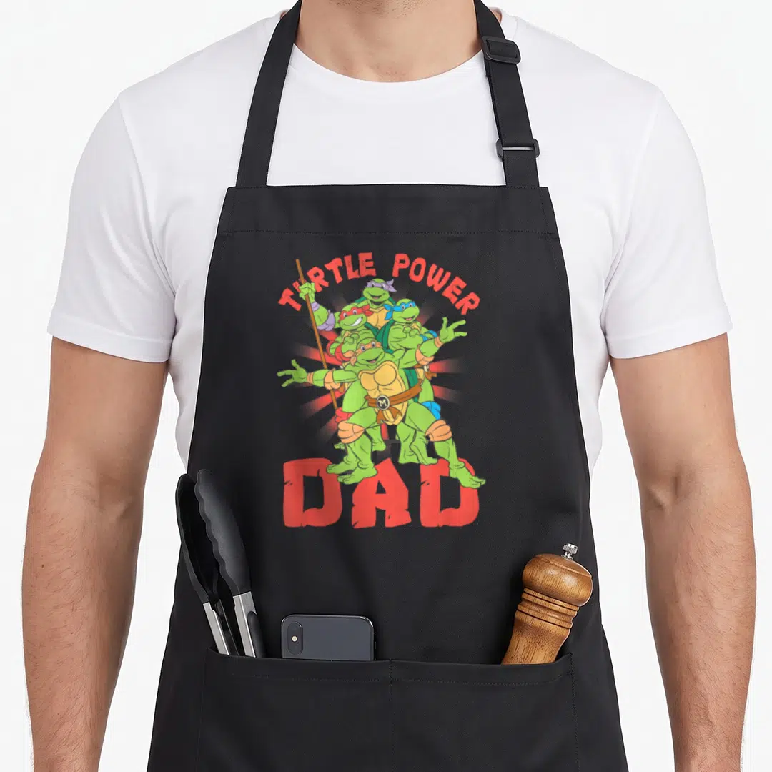 Power turtle dad apron - Four stacked green turtles with red TURTLE POWER DAD text and brown belt — shown on a black cotton canvas apron against a white background
