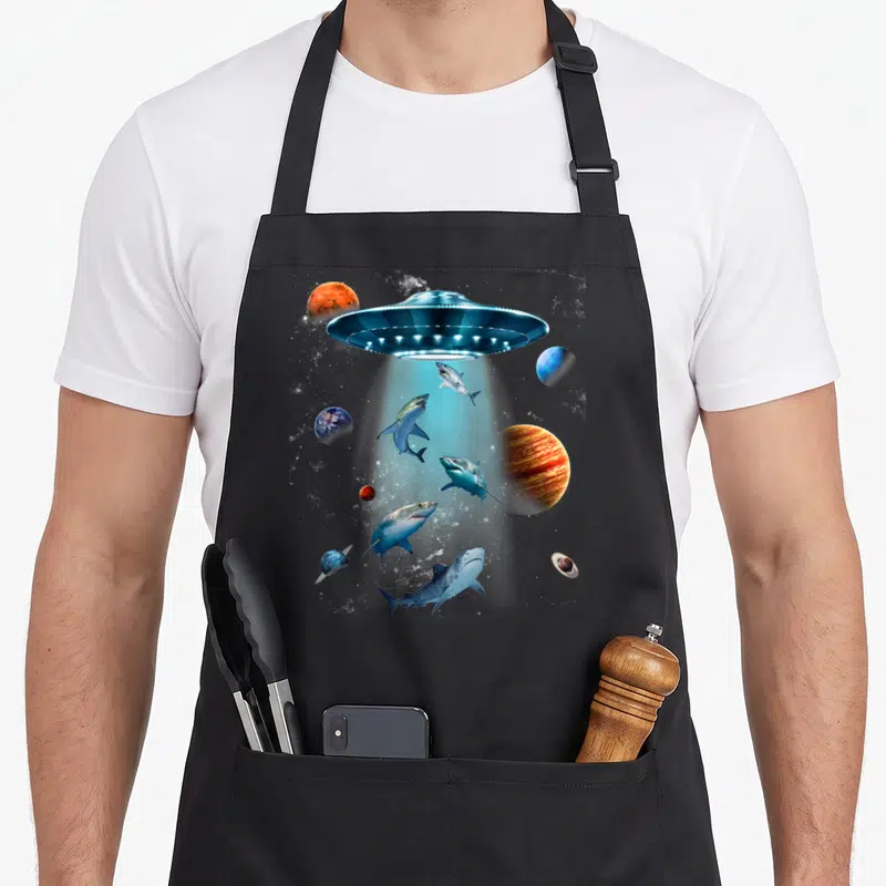 Galaxy fish apron - UFO beaming up five sharks among orange and blue planets in space — shown on a black cotton canvas apron against a white background