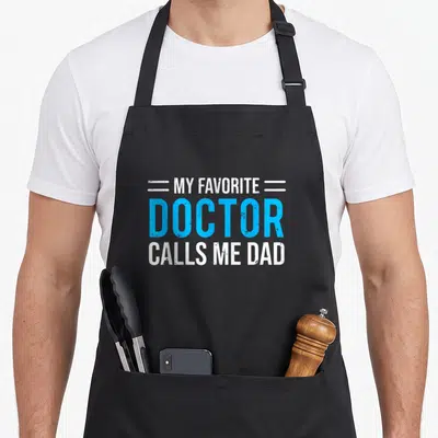 Call doctor with apron - White and blue stacked text reading My Favorite Doctor Calls Me Dad — shown on a black cotton canvas apron against a white background