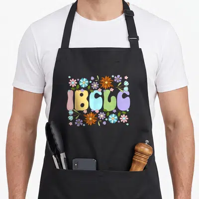 Lactation consultant apron - IBCLC bubble text surrounded by pink, yellow, blue, green, purple, and orange cartoon — shown on a black cotton canvas apron against a white background