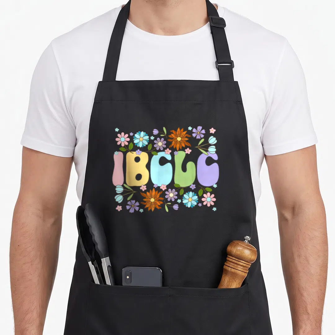 Lactation consultant apron - IBCLC bubble text surrounded by pink, yellow, blue, green, purple, and orange cartoon — shown on a black cotton canvas apron against a white background
