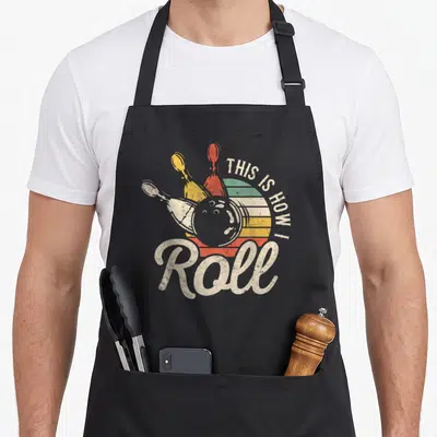 Bowling roll apron - Retro bowling ball and pins over striped sun with off-white text — shown on a black cotton canvas apron against a white background