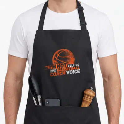 Yelling basketball coach apron - Orange basketball above stacked orange and white text reading I'm not yelling this is — shown on a black cotton canvas apron against a white background