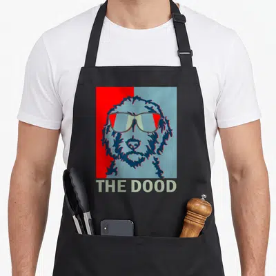 Dood doodle goldendoodle apron - Cartoon dog face with sunglasses on split red and blue background with text THE DOOD — shown on a black cotton canvas apron against a white background