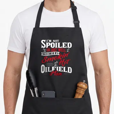 Oilman wife apron - Stacked text I'M NOT SPOILED I'M JUST WELL TAKEN CARE OF BY A Smokin' Hot OILFIELD Man with three — shown on a black cotton canvas apron against a white background