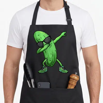 Dancing pickle apron - Green cartoon cucumber dabbing in black sunglasses and sneakers — shown on a black cotton canvas apron against a white background