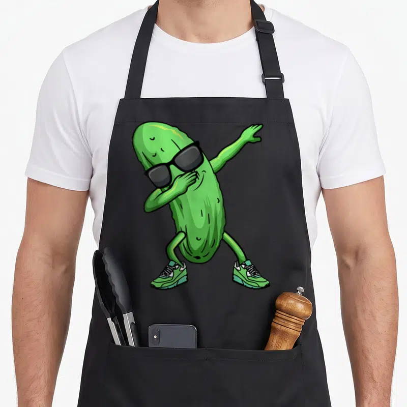 Dancing pickle apron - Green cartoon cucumber dabbing in black sunglasses and sneakers — shown on a black cotton canvas apron against a white background