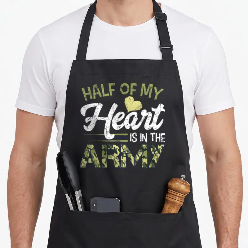 Vintage army wife apron - Stacked text HALF OF MY Heart IS IN THE ARMY with camo fill and distressed olive green accents — shown on a black cotton canvas apron against a white background