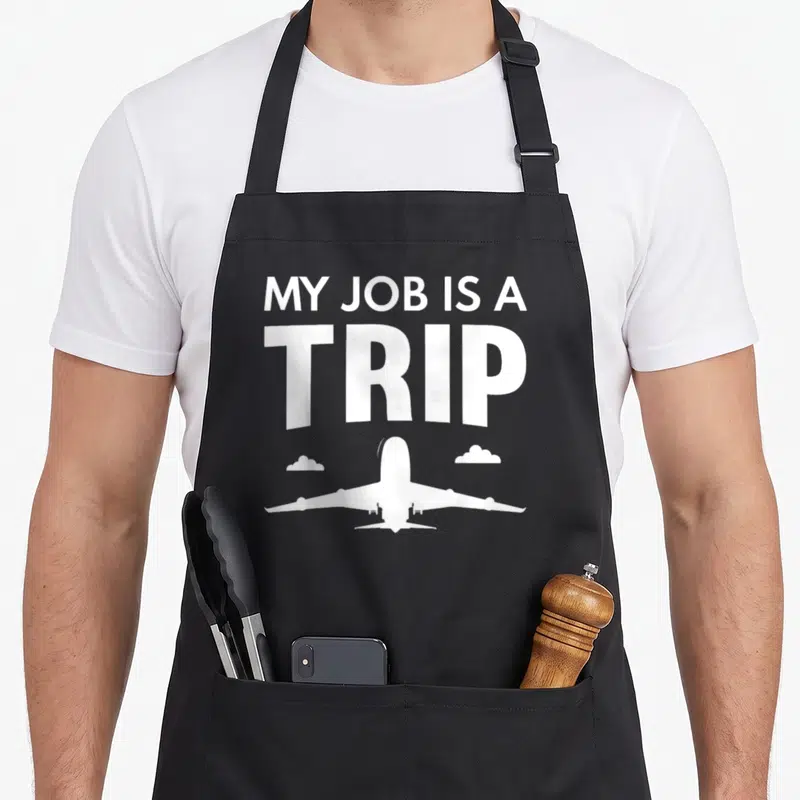 Flight travel aprons - White stacked text MY JOB IS A TRIP above airplane silhouette and two small clouds — shown on a black cotton canvas apron against a white background