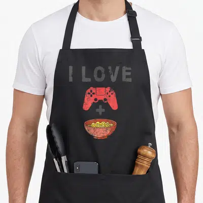 Ramen gaming apron - I LOVE text above red controller plus yellow noodles in bowl — shown on a black cotton canvas apron against a white background
