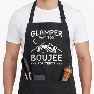 Glamping luxury apron - White distressed Glampers Way Too Boujee For Tents text above mountains with moon stars — shown on a black cotton canvas apron against a white background