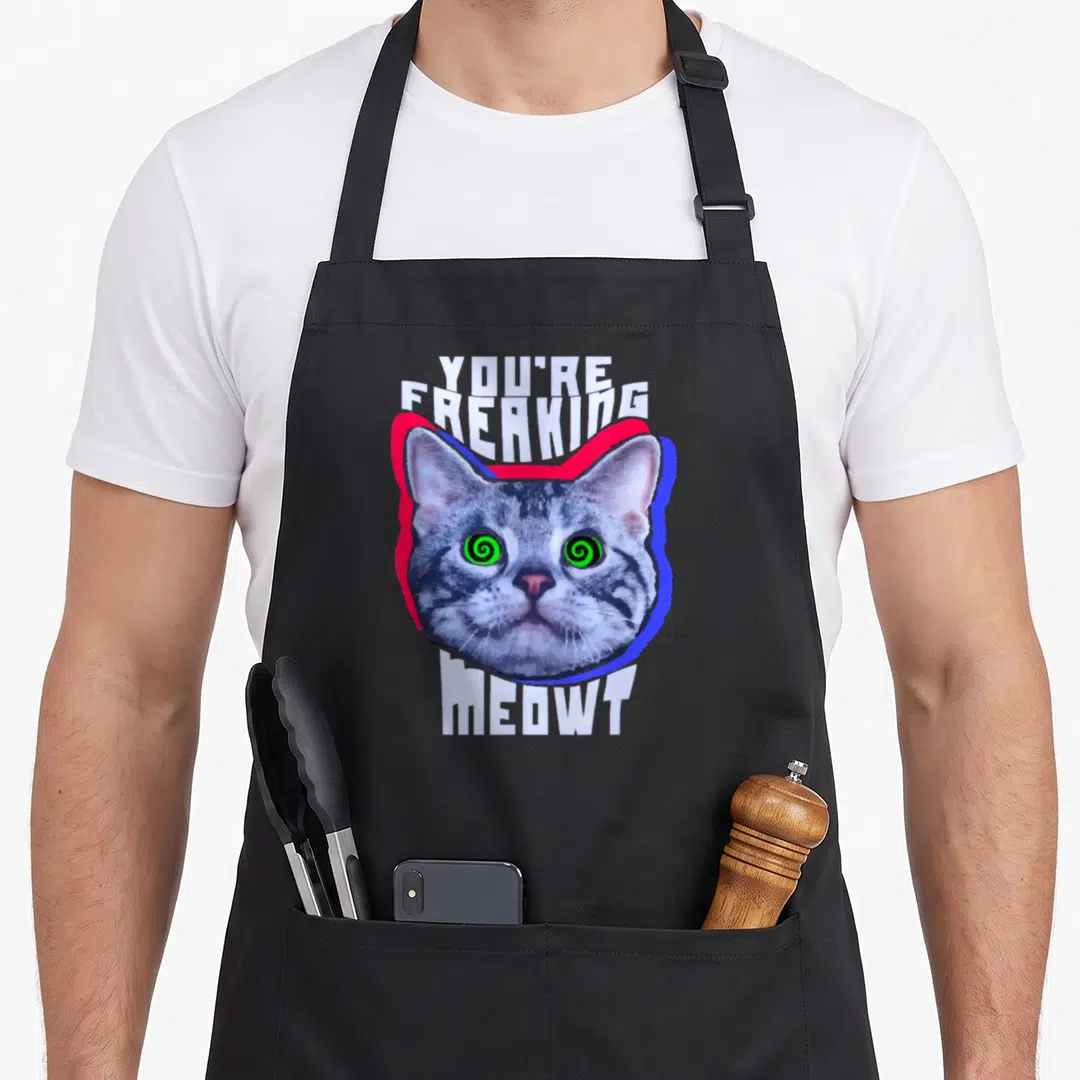 Freaking kitty cat apron - Grey cat head with green spiral eyes and white text YOU'RE FREAKING MEOWT over red and blue — shown on a black cotton canvas apron against a white background