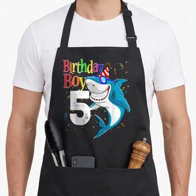 Shark birthday apron - Cartoon blue shark with sunglasses and party hat beside Birthday Boy text and number 5 — shown on a black cotton canvas apron against a white background
