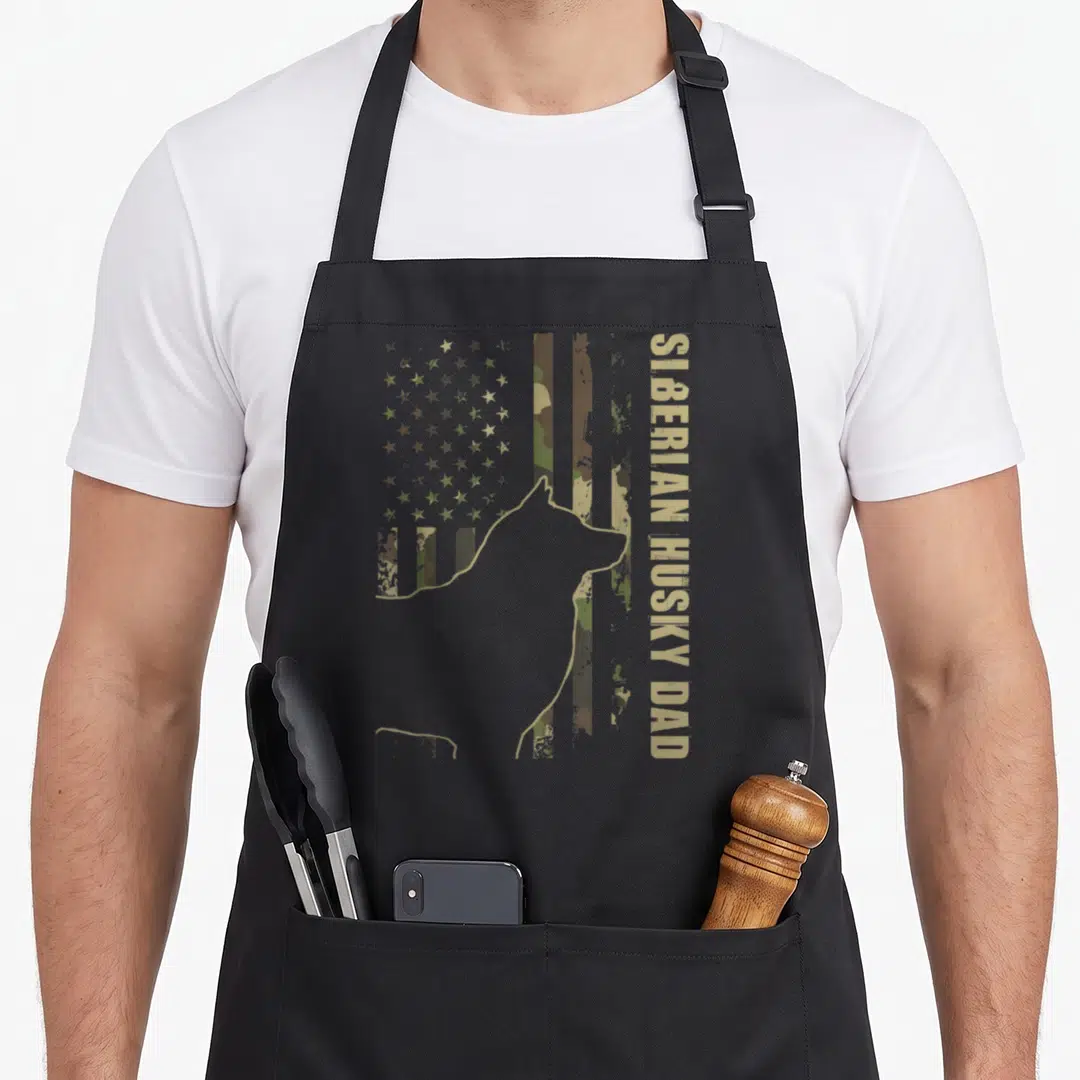 Siberian dog trainer apron - Grey husky silhouette over distressed camo flag with vertical tan text SIBERIAN HUSKY DAD — shown on a black cotton canvas apron against a white background