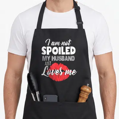Wife husband apron - I am not SPOILED MY HUSBAND JUST Loves me text with red lips — shown on a black cotton canvas apron against a white background