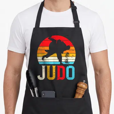 Throw judo addict apron - Judo practitioner silhouette against striped retro sun with kanji and JUDO text — shown on a black cotton canvas apron against a white background