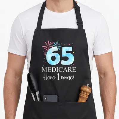 65th come and get it apron - Blue 65 with MEDICARE Here I come! text and pink white yellow fireworks stars — shown on a black cotton canvas apron against a white background