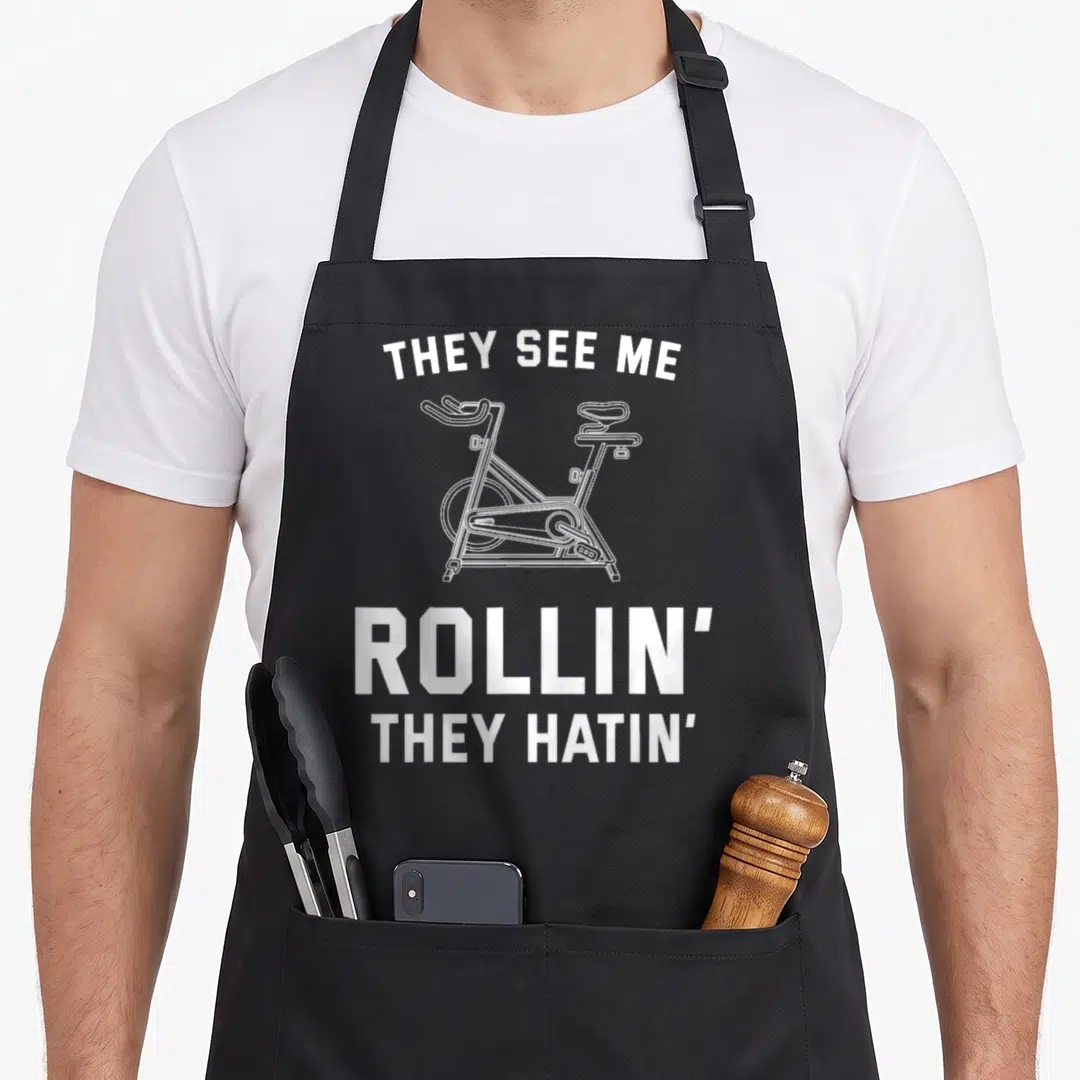 Spin see me aprons - White line art stationary bike between THEY SEE ME ROLLIN THEY HATIN text — shown on a black cotton canvas apron against a white background