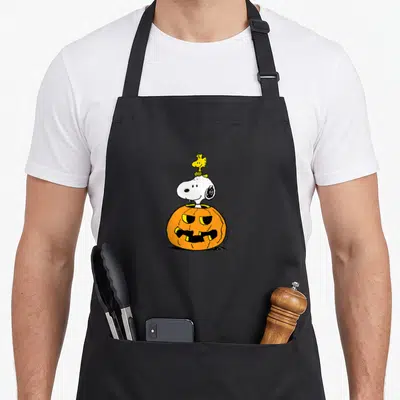 Funny woodstock halloween kitchen aprons - Snoopy head emerging from orange pumpkin with yellow Woodstock perched — shown on a black cotton canvas apron against a white background