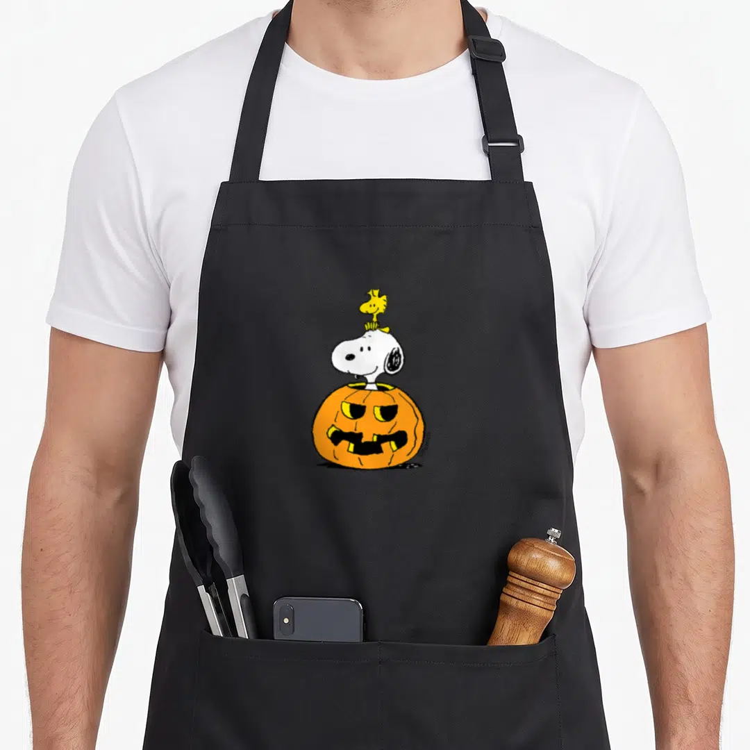 Funny woodstock halloween kitchen aprons - Snoopy head emerging from orange pumpkin with yellow Woodstock perched — shown on a black cotton canvas apron against a white background