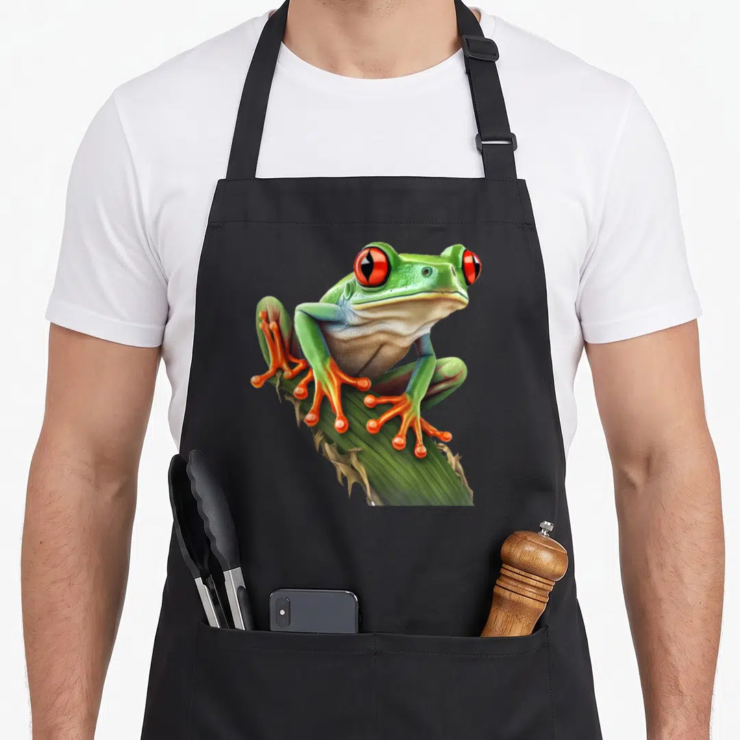 Rainforest tree apron - Realistic green frog with red eyes and orange feet perched on a diagonal leaf — shown on a black cotton canvas apron against a white background