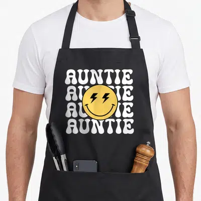 Auntie custom face apron - Yellow smiley face with black lightning bolt eyes over four stacked white AUNTIE text lines — shown on a black cotton canvas apron against a white background