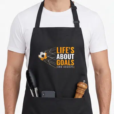 Goal boy soccer apron - Soccer ball hitting net beside stacked orange and white text reading Life's About Goals — shown on a black cotton canvas apron against a white background