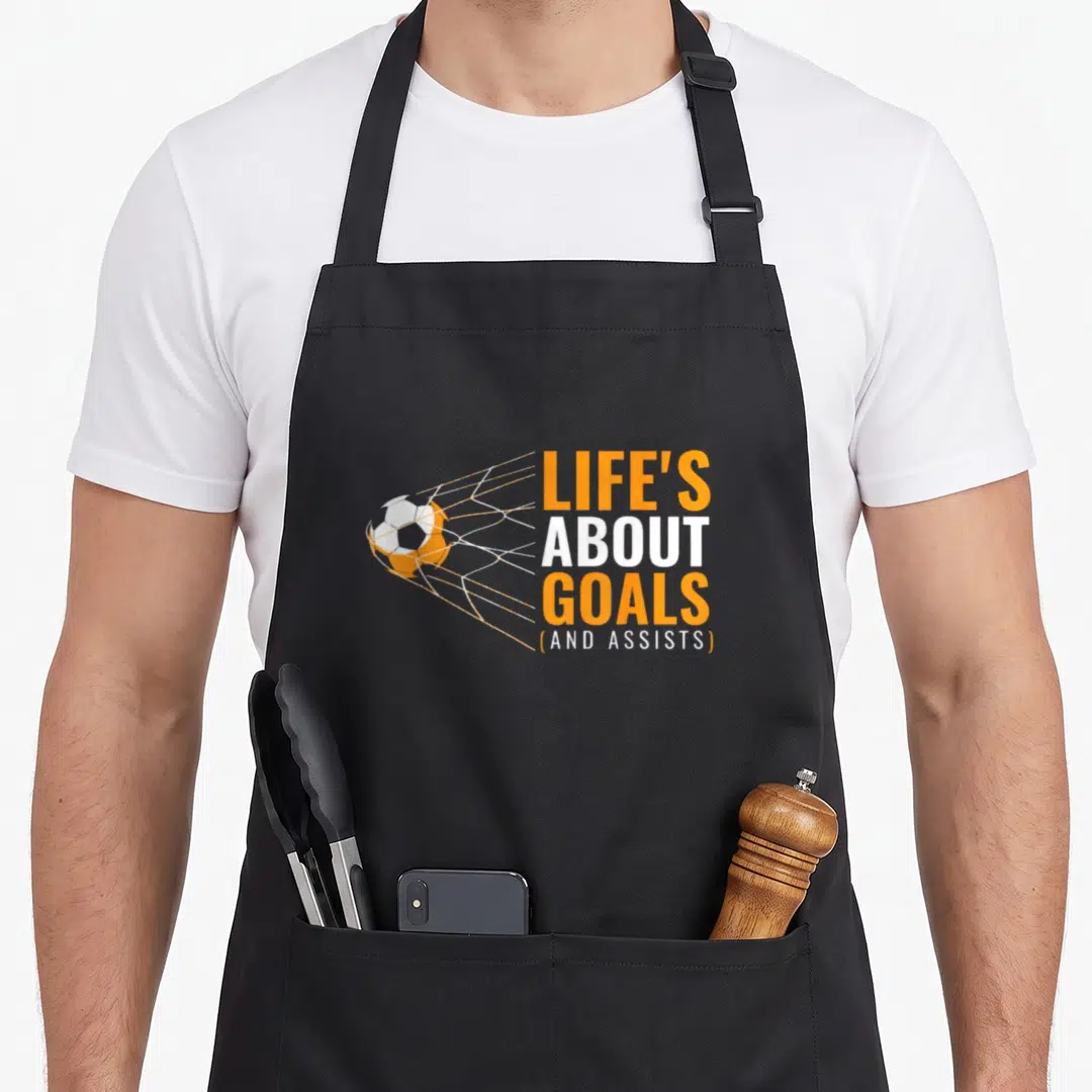Goal boy soccer apron - Soccer ball hitting net beside stacked orange and white text reading Life's About Goals — shown on a black cotton canvas apron against a white background