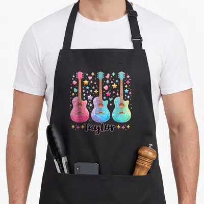 Groovy 80s apron - Three pink blue teal yellow and black guitars with scattered stars hearts and Taylor script — shown on a black cotton canvas apron against a white background