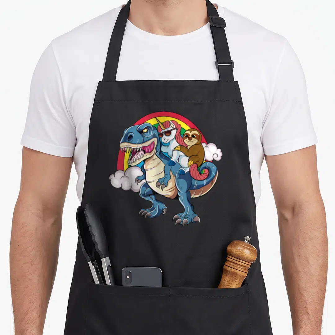 Magical dinosaur apron - Blue dinosaur carries pink-maned unicorn and brown sloth under rainbow with clouds — shown on a black cotton canvas apron against a white background