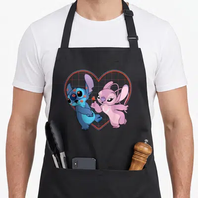 Kiss angels in aprons - Blue cartoon character with red lips next to pink character inside red grid heart — shown on a black cotton canvas apron against a white background