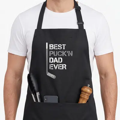 Puckn father day apron - White distressed hockey stick beside stacked text reading Best Puck'n Dad Ever — shown on a black cotton canvas apron against a white background
