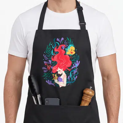 Ariel flounder mermaid apron - Cartoon Ariel with red hair, Flounder, Sebastian, teal seaweed, purple coral, and yellow — shown on a black cotton canvas apron against a white background