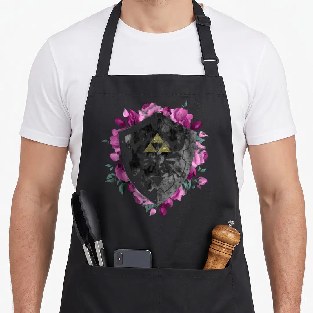 Force floral apron - Grey shield with gold triforce surrounded by pink roses and green leaves — shown on a black cotton canvas apron against a white background