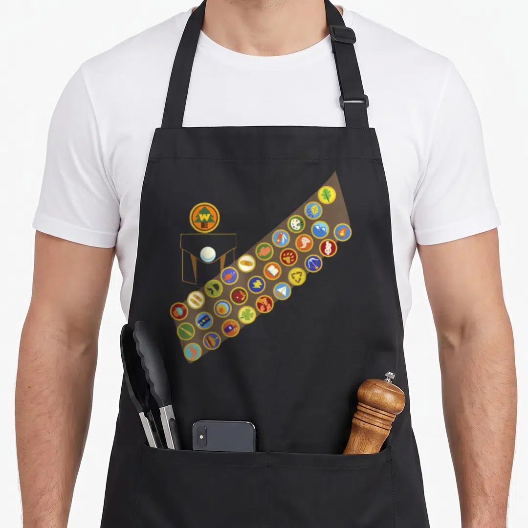 Russell halloween apron - Cartoon sash with circular merit badges showing archery, cooking, and first aid symbols — shown on a black cotton canvas apron against a white background