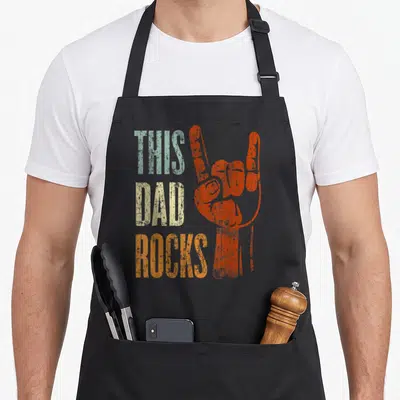 Father heavy metal apron - Stacked THIS DAD ROCKS text next to orange rock hand sign in distressed blue and beige colors — shown on a black cotton canvas apron against a white background