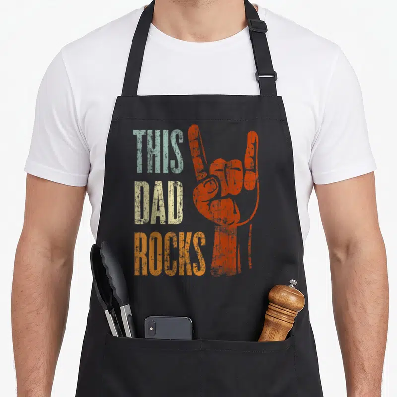 Father heavy metal apron - Stacked THIS DAD ROCKS text next to orange rock hand sign in distressed blue and beige colors — shown on a black cotton canvas apron against a white background