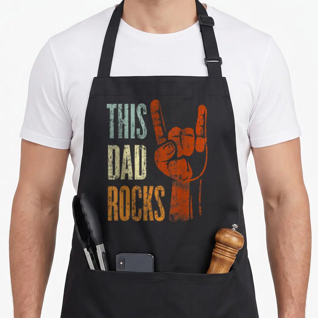 Father heavy metal apron - Stacked THIS DAD ROCKS text next to orange rock hand sign in distressed blue and beige colors — shown on a black cotton canvas apron against a white background