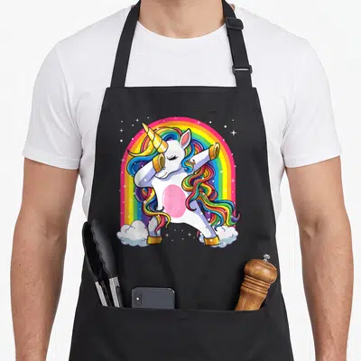 Cute dabbing rainbow apron - Cartoon white unicorn dabbing with gold horn and rainbow mane under a rainbow arch — shown on a black cotton canvas apron against a white background