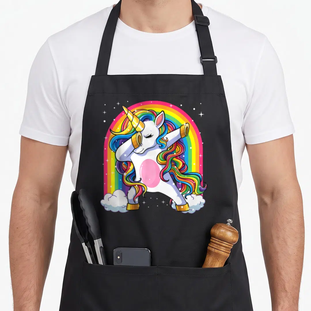 Cute dabbing rainbow apron - Cartoon white unicorn dabbing with gold horn and rainbow mane under a rainbow arch — shown on a black cotton canvas apron against a white background