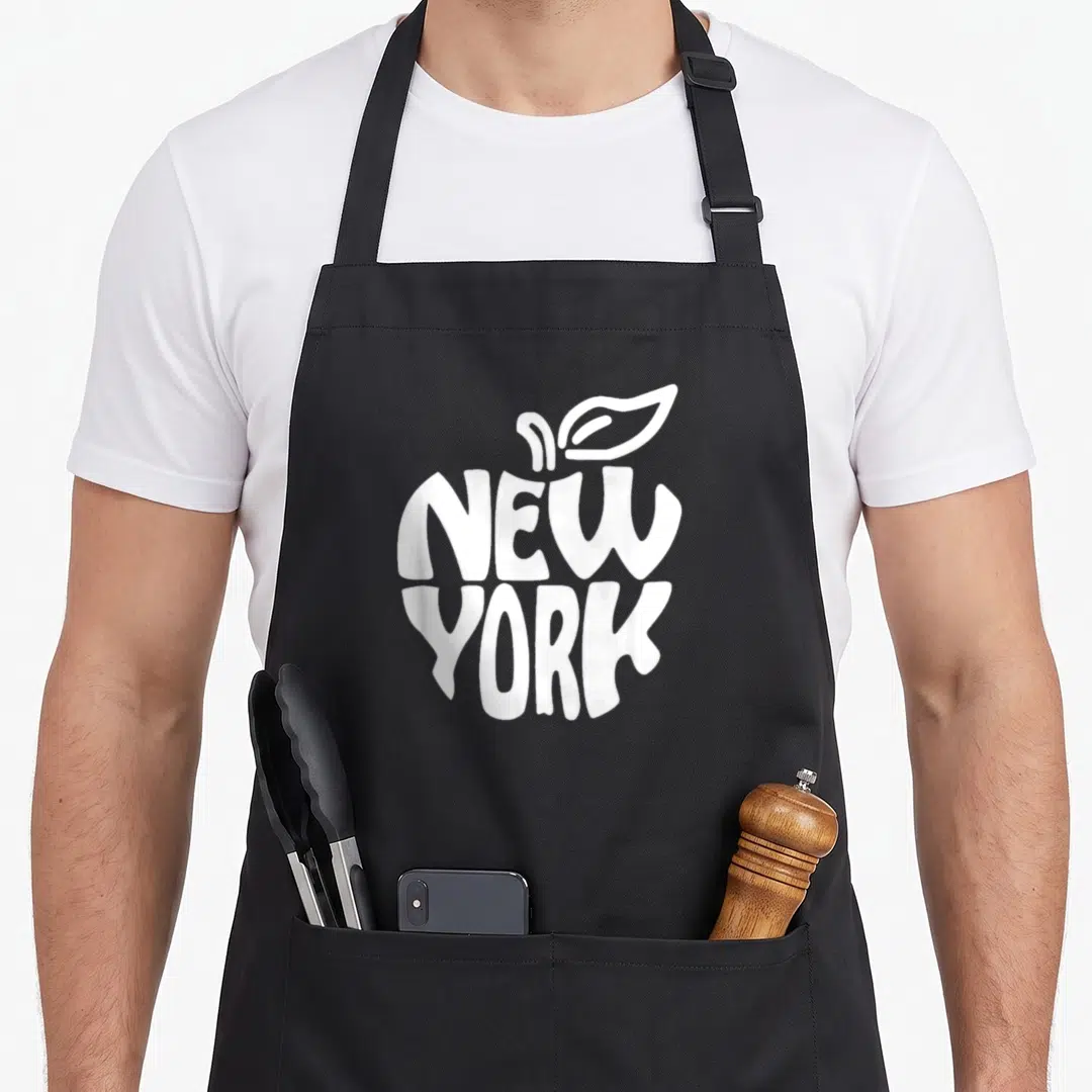 Funny nyc new york apron - White NEW YORK text shaped like an apple with leaf and stem on black fabric — shown on a black cotton canvas apron against a white background