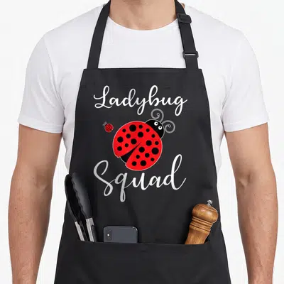 Cute squad ladybug apron - Red cartoon ladybug with black spots and white script text reading Ladybug Squad — shown on a black cotton canvas apron against a white background
