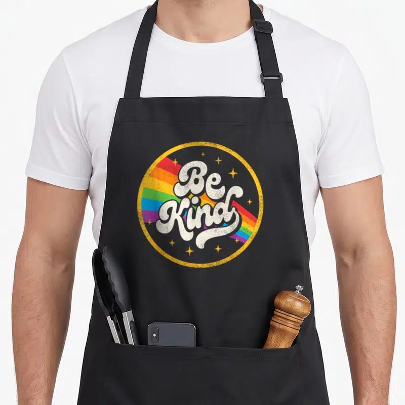 Lgbtq kind lgbt ally gay pride apron - Retro Be Kind text over rainbow stripes inside yellow circle with stars — shown on a black cotton canvas apron against a white background